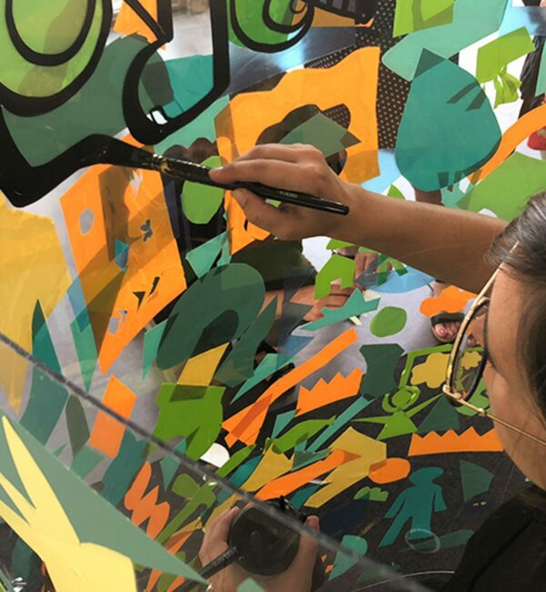 une artiste à lunettes rondes dessine à la ligne noire autour de formes de couleurs sur un support transparent plexi art en animation fresque géante lors d'un team building
