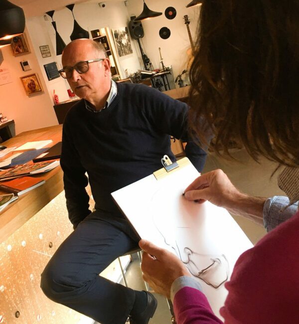 un artiste aux cheveux longs et veste fuchsia en train de réaliser la caricature noir et blanc format A3 en face de son modèle homme assis en face de lui en animation caricature papier à Lyon