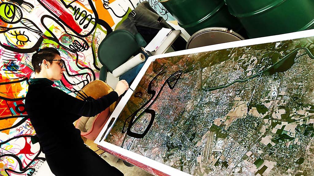 l'artiste aNa dans son atelier de Lyon penchée sur sa table en train de peindre une fresque sur plexi à partir du plan google earth imprimé de la Ville de Lyon métropole