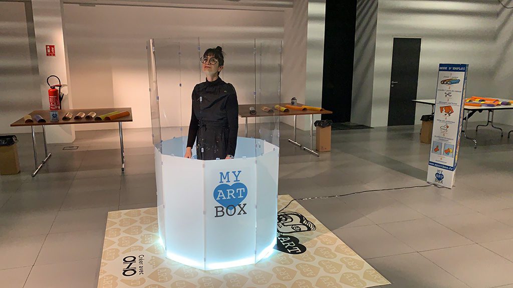Une artiste se tient debout dans un tube cylindrique composée d'une partie haute totalement transparente et d'une partie basse blanche rétro éclairée et présentant le logo my art box bleu et gris