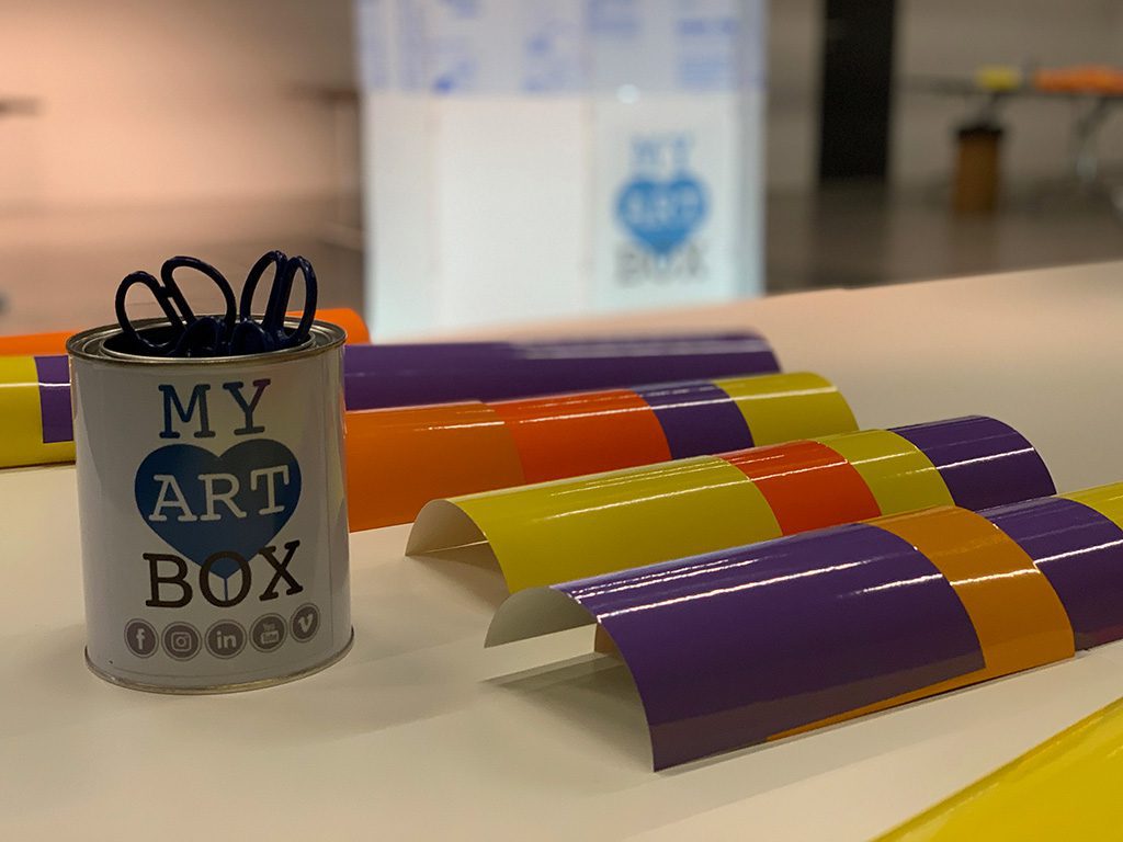 matériel du protocole artistique d'atelier team building myartbox composé d'un pot contenant des ciseaux de découpage, des adhésifs de couleurs posés sur une table, et dans le fond le support lumineux de fresque plexi lumineuse de l'artiste aNa