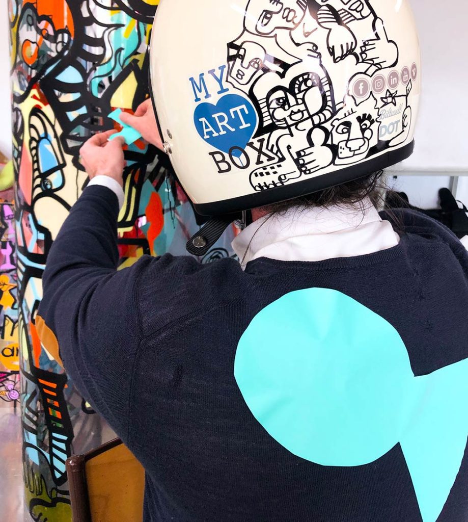 un homme qui porte un casque blanc décoré par l'artiste aNa pour myartbox en pull bleu avec un poisson d'avril géant vert collé dans le dos