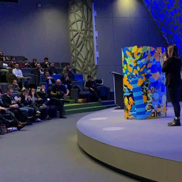 Une personne sur scène devant un publique en assemblée présente le résultat d'une animation Convention Animation Fresque Totem Box XL Tube plexi My Art Box
