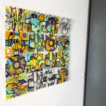 Un tableau DIY Happiness management my art box composé de 16 plaques plexi personnalisée avec des couleurs vives et posé sur un mur blanc à coté d'un mur gris