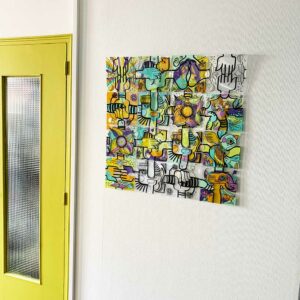 Tableau décoratif composé de plaques de plexiglass dessinées par l'artiste aNa et associées pour former une composition originale sur un mur blanc à coté d'une porte de séparation verte