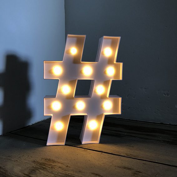 Un hashtag blanc lumineux posé sur un planché en bois dans un angle de murs blancs pour illustrer l'importance de bien choisir son hashtag afin de répondre à la question de comment développer son business avec instagram