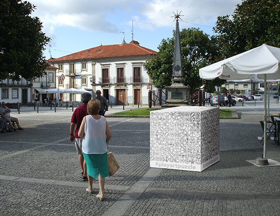 Un homme en polo bordeaux et une femme top blanc jupe verte passent devant un cube dessiné en noir blanc et comportant le hashtag Play Art Puzzle au milieu d'une place publique pavée