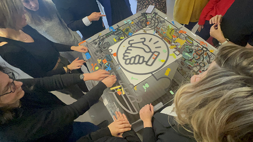 La Fresque de la Considération à Saint Etienne avec 100 collaborateurs 2 Les participantes lors de la fresque de la considération My Art Box à Saint Etienne sont massées autour du plexi pour coller leurs idées personnelles