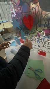 Un participant à une fresque hybride à Paris colle son découpage de vinyle bleu représentant un voilier sur le plexi collaboratif pré-imprimé avec le dessin digital de aNa artiste
