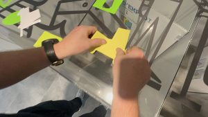 Un participant à l'animation fresque anniversaire entreprise à Genève colle sur le support plat en plexi My ARt Box un découpage de charriot élévateur découpé dans du vinyle jaune