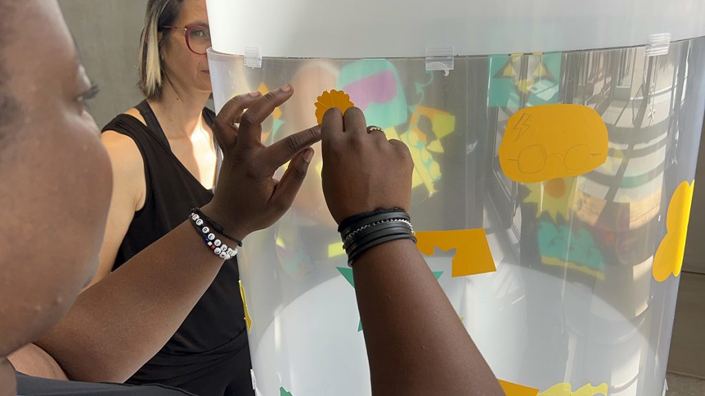 Une participantes colle son idée sur le cylindre en plexiglas de l'artiste aNa lors de l'animation fresque éco-responsable à Genève