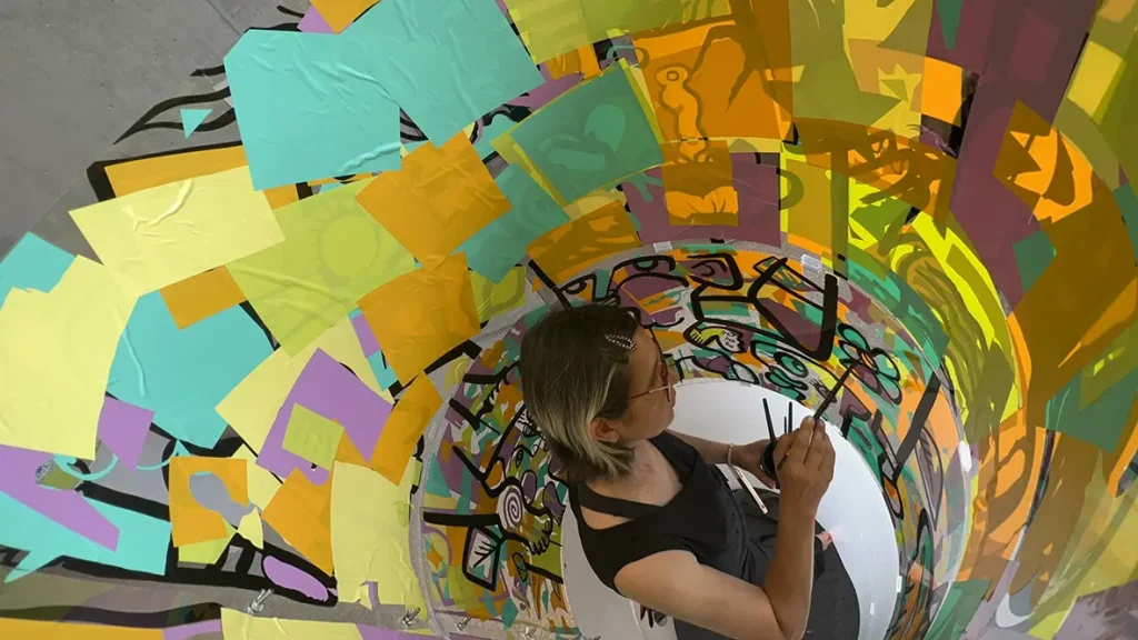 Vue en plongée de l'artiste aNa peignant une fresque collaborative à l'intérieur d'un tube transparent lors d'un événement à la Comédie de Genève.