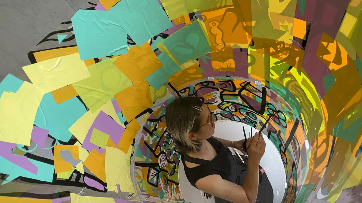 Vue en plongée de l'artiste aNa peignant une fresque collaborative à l'intérieur d'un tube transparent lors d'un événement à la Comédie de Genève.