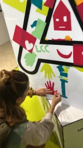 Participant en atelier sans peinture appliquant des formes colorées sur une sculpture collaborative en plexiglas (protocole Anthropotekhnè) My Art Box.