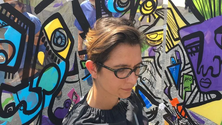 Artiste avec lunettes peignant une fresque colorée sur un support transparent devant un public