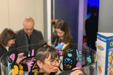 Une artiste peint à l'intérieur d'un grand cylindre transparent entourée de participants lors d'une pose de première pierre.