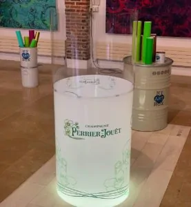 Un tube oeuvre commune de l'artiste aNa et MYARTBOX vide avant le début de l'activité team building avec tout le matériel nécessaire à l'activité dans le fond posé sur un baril de 200 l avec logo myartbox et au premier plan le Totem plexi art en deux parties une haute totalement transparente et la base blanche rétro éclairée et présentant le logo vert de la maison de Champagne Perrier Jouet