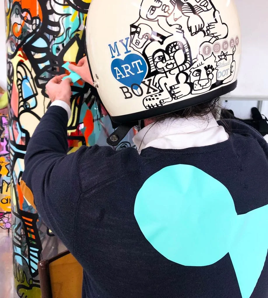 un homme qui porte un casque blanc décoré par l'artiste aNa pour myartbox en pull bleu avec un poisson d'avril géant vert collé dans le dos