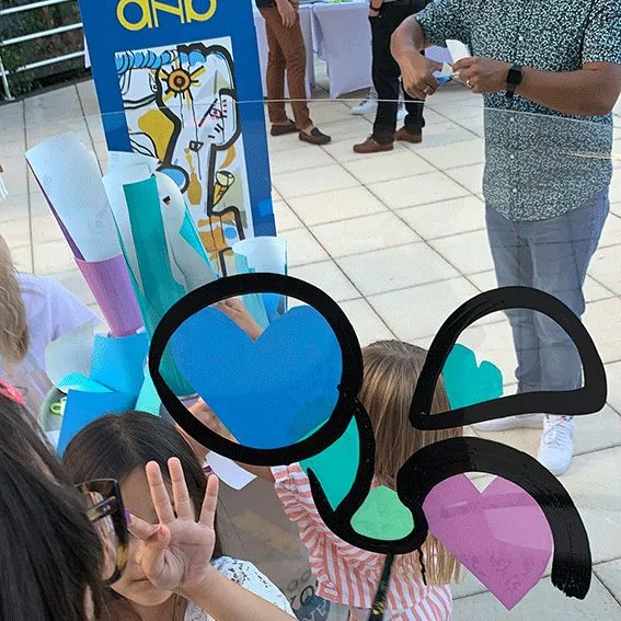 Plusieurs mains d'invités coloriant ensemble une fresque artistique sur plexiglas lors d'une fête d'anniversaire.
