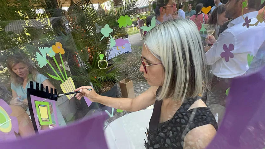 ana artiste à l'intérieur de son tube fresque en garden party avec les participants autour créant en même temps des adhésifs découpés qu'ils collent sur le support transparent de l'artiste