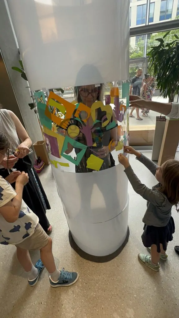 ana artiste en train de peindre dans son tube en plexiglas et des participantes en animation fresque éco-responsable à Genève