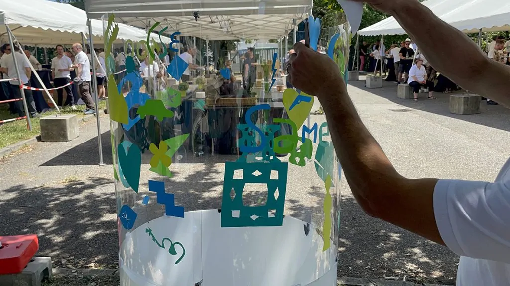 L'animation originale éco-responsable fresque en garden party à Grenoble réalisée par aNa commence par la participation des collaborateurs qui collent des formes découpées dans du vinyle coloré sur le tube plexiglas de l'artiste